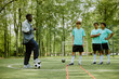 © Seventyfour - Black man coach instructing three teenage boys on soccer field during training session, teenagers standing attentively in sportswear, soccer goal and trees visible in background