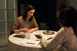 © Mediaphotos - Caucasian young woman sitting at round table reading tarot cards for Black young woman, discussing predictions and meanings, both engaging in conversation with notebook and candle on table