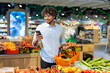 © Liubomir - A young man browsing his smartphone while selecting fresh vegetables in a supermarket. He holds an orange basket with groceries as he explores the store's produce section amidst colorful displays.