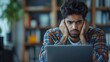 © FANG - Stressed and frustrated young Indian man with hands framing his face, dark curly hair and beard, furrowed brows, wearing blue-orange-white checkered shirt over white T-shirt, with a dark laptop screen