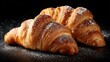 © Lebah - Two golden-brown croissants, dusted with powdered sugar, rest on a dark surface