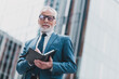 © deagreez - Confident senior businessman holding notebook outdoors near a modern office building, representing professionalism and success