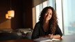 © joniex - A smiling woman with curly hair sits at a table by a window, engaging with a book in a well-lit, modern interior.