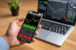 © olgasun - Hand holding phone displaying financial charts, laptop, digital trading.