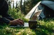 © Marina Demidiuk - Close-up of hands typing on a laptop, sitting on the grass near a tent in a forest. Man is working outdoors using a computer. Close-up of a man using a notebook and drinking coffee, camping in nature.