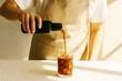 © Sergey - Barista in apron pouring cold brew coffee from black bottle into glass with ice, warm sunlight, minimal indoor background, morning beverage concept