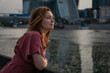 © Sofia - Woman in a red dress leaning on a railing, looking thoughtfully across the river with Moscow City skyscrapers blurred in the background