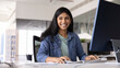 © fizkes - Happy young Indian professional woman posing at office workplace, sitting at computer monitor, working on Internet startup project, looking at camera, smiling, laughing. Banner shot