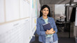 © fizkes - Positive young Indian company director woman in casual denim clothes posing at white board in office workspace, standing with hands folded, looking at camera with friendly smile. Banner shot