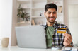 © Prostock-studio - Easy Payment And Cashback Concept. Portrait of excited millennial guy holding credit card in hand and using pc sitting at table at home, ordering purchases in web store making financial transaction