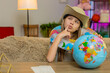 © Andrii Iemelianenko - Young child girl sitting at home table wears a travel hat and inflatable ring while looking at a globe, imagining a beach vacation. School kid enjoys dreaming about travel planning a seaside holiday