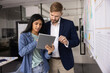 © fizkes - Diverse couple of young business colleagues using online application for project management tablet computer, talking at scrum board with sticky notes, discussing project plan, tasks scheduling