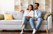 © Prostock-studio - Joyful Indian couple, comfortably sitting on a sofa in their homey space, sharing a light, dreamy moment while gazing aside and sharing tender smiles, free space