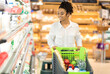 © Prostock-studio - African woman on grocery shopping in supermarket pushing cart walking along shelves buying food and goods in store. Female customer choosing groceries in local shop. Empty space