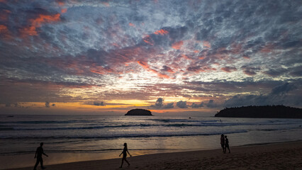 Wall Mural - Golden Embrace above Kata beach.
The setting sun paints the horizon with golden light, reflecting across gentle waves as silhouettes of beachgoers stroll peacefully along the shoreline.