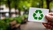 © Анна Терелюк - Hand holding recycling symbol in park. A person holds a card with a green recycling symbol in a lush park setting surrounded by trees and benches.