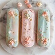 Three exquisite eclairs, glazed in pastel hues and embellished with gold flakes and delicate fondant flowers, sit beautifully on a marble serving plate, inviting a sweet indulgence