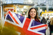 Cheerful young girl traveling in United Kingdom during Christmas holidays, posing at traditional street fair with national British flag in hands