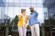 © sofiko14 - Two people wearing VR headsets in front of a modern building explore a virtual environment together, one holding a tablet.