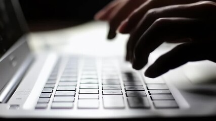 Hands type on a backlit keyboard of a laptop in shadow, close up angle shot