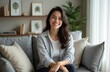 © Maryna - Smiling woman in casual attire sits on gray couch in cozy living room. Long, dark hair, relaxed, confident expression. Interior features neutral tones with artwork, plants, creating comfortable,