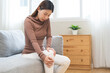 © KMPZZZ - Health care of knee pain concept, suffering asian young woman arthritis sitting joint ache, sore cramp or sprain tendon in leg on sofa at home. Osteoarthritis chronic disease of body problem, recovery
