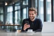 © Yuliia Malovana - A smiling businessman in black shirt with a laptop using his smartphone in coworking space, with coffee cup and modern interior in the background. A man smiles while looking at his phone at a cafe.