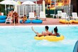 © Damian - Young adults relax together, floating in a yellow tube on a lazy river ride at the park, with other people sunning and enjoying the day around the blue waters
