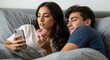 © Manji - Young Couple Relaxing in Bed Enjoying Donut and Smartphone