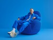© Olesia Bobrus - Fashionable man dressed in blue clothing and beige sneakers, sitting comfortably on a blue armchair in a vibrant blue monochrome room, smiling brightly while looking at the camera
