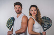 © EricMiguel - Confident male and female padel tennis players posing back to back with rackets in white sportswear against plain background