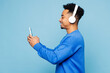 © ViDi Studio - Side view young man of African American ethnicity wear sweatshirt casual clothes listen to music in headphones use mobile cell phone isolated on plain pastel light blue background. Lifestyle concept.