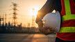 © sabrang - Close-up of an electrical engineer holding a white hard hat, with a power plant and transmission towers in the background at sunset.