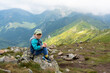 © Maryia - Happy young boy in cap and blue jacket sitting on rocky mountain boulder enjoying spectacular alpine valley views on clear day. Concept of family hiking achievements and childhood mountain experiences