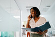 © djile - Smiling black businesswoman talking on phone and holding folder in office corridor