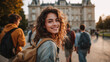 © Dirk - Group of young people with backpacks, smiling towards the camera in front of university building on campus. Woman is looking from behind her shoulder, carrying a canvas bag. University students.