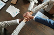 © pressmaster - Diverse group of young adult and middle aged men and women stacking hands in teamwork gesture on wooden table, business documents and charts surrounding collaborative action