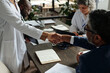 © pressmaster - Caucasian woman in lab coat shaking hands with Black middle aged man in suit while two other professionals working at table with documents and laptop