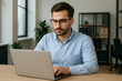 ©  JAMAL PRODUCTION - Focused young businessman in glasses working diligently on his laptop in a bright, modern office setting, embodying professionalism and remote productivity.