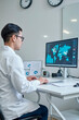 © AnnaStills - Asian young adult man sitting at desk analyzing global data on large computer monitor in modern office, wearing lab coat and working with statistical charts and world map graphics