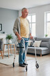 © AnnaStills - Senior Caucasian man standing in living room using walker for support, looking forward with neutral expression, natural light coming through windows, casual home environment