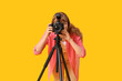 © Pixel-Shot - Young woman taking photo with modern camera on tripod on yellow background