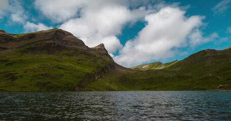 Wall Mural - Alpine lake surrounded by lush green slopes and dramatic rocky peaks partially covered with clouds, under a bright blue sky