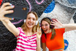 © TrueShot | Westend61 - Happy female friends taking selfie through smart phone while gesturing against graffiti wall