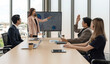 © chadchai - Asian businessman in suit raise his hand to ask a question to presenter standing in front of a large digital monitor. Business executives team meeting in modern office.
