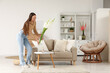 © Pixel-Shot - Young woman putting vase with gladiolus flowers on table in living room