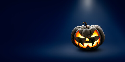 Wall Mural - A lit scary halloween lit Jack O Lantern at night under the glow of a spotlight on a wide dark blue background with copy space.