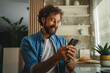 © Miljan Živković - Smiling man using smart phone in kitchen reading good news