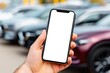 © Cheewynn - A hand holds a smartphone with a blank white screen in front of parked cars in an outdoor setting