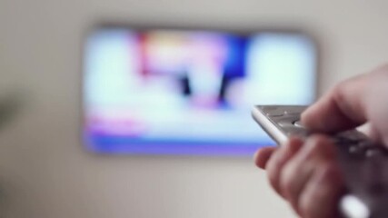 Close-up of a hand holding a TV remote control, with a blurry TV screen in the background